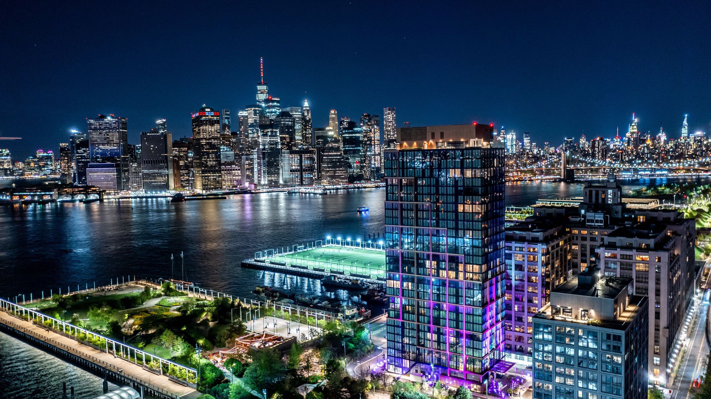 Quay Tower lit at night above Brooklyn Bridge Park, with the Pier 5 fields and lower Manhattan beyond